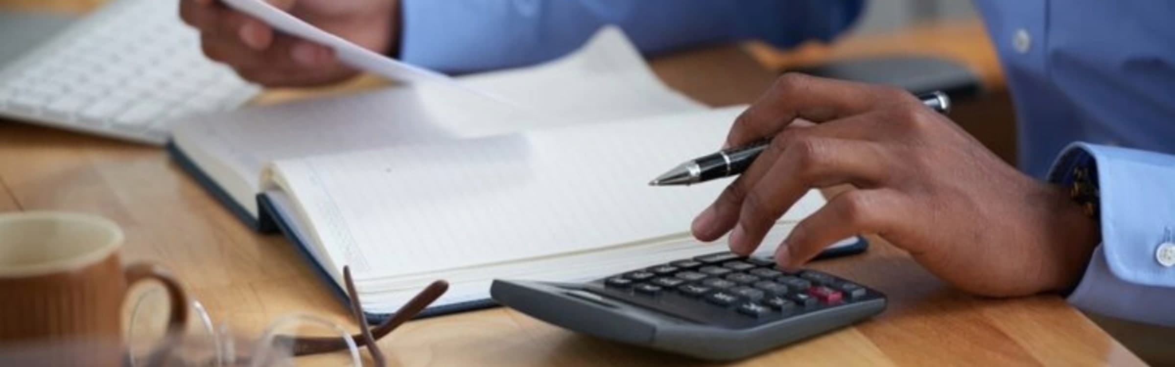A hand working through figures on a desk calculator beside an open ruled notebook, a pen, reading glasses and a mug.