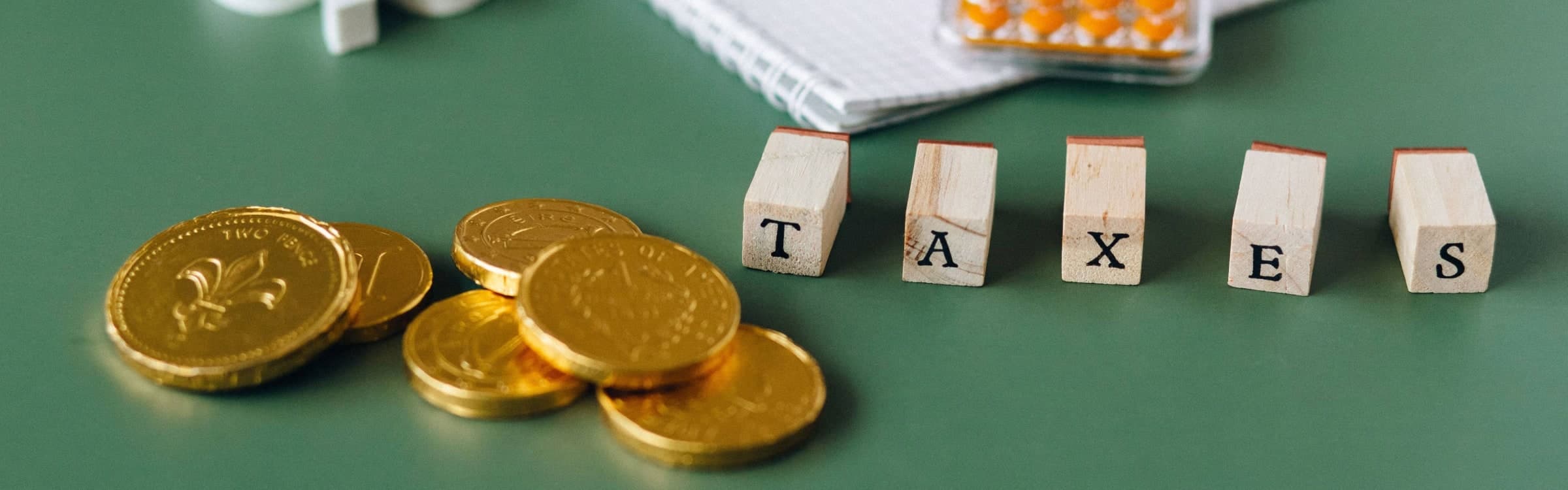 Gold coins stacked on a green desk beside wooden letter blocks spelling the word TAXES, with a spiral notebook behind them.